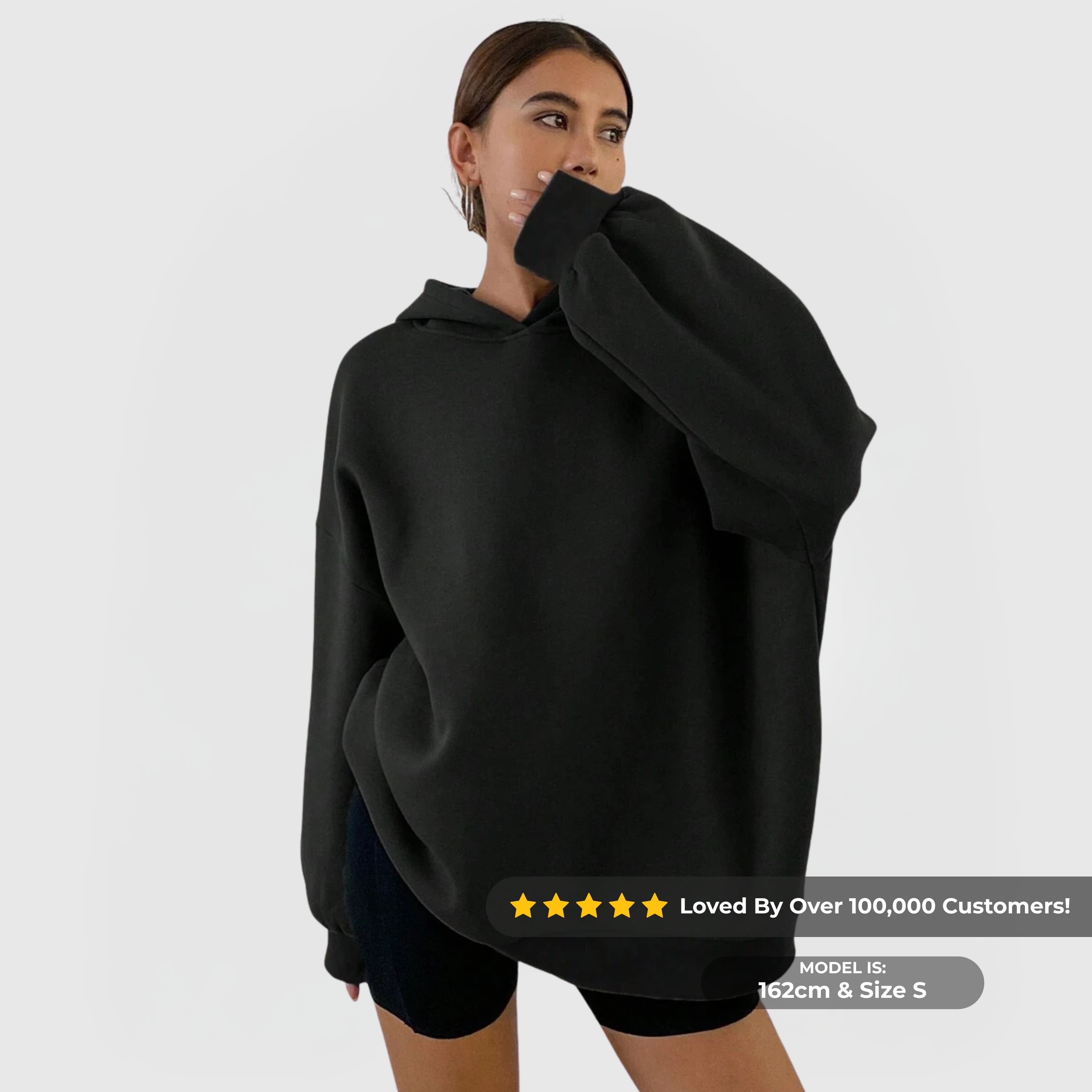 Woman wearing black oversized essential hoodie with dropped shoulders and kangaroo pocket, posing against plain background.