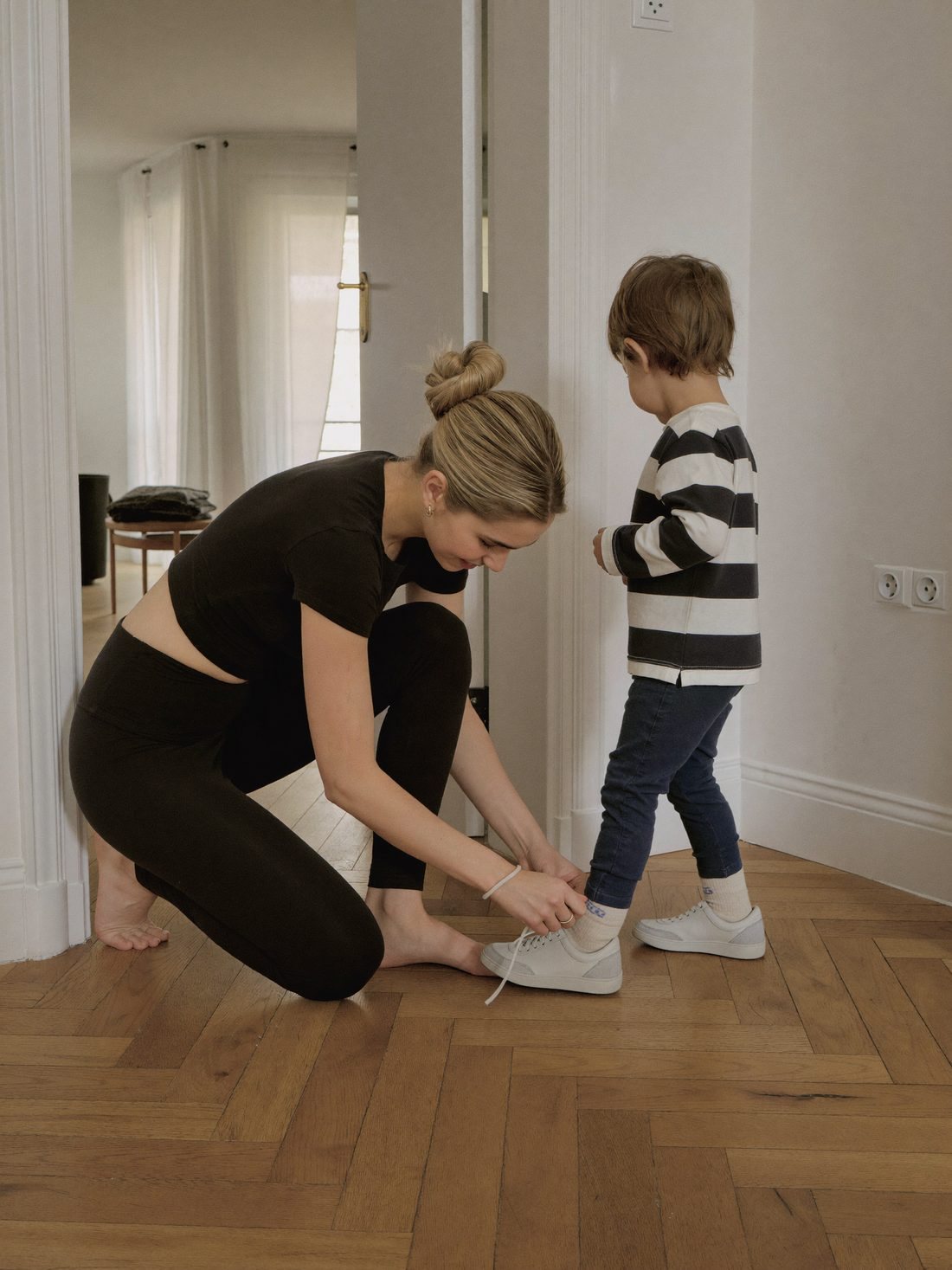 Nainen kyykistyy solmimaan lapsen kengännauhat legginsseissä